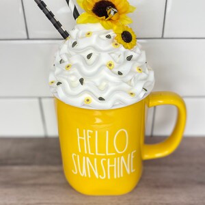 May include: Yellow ceramic mug with the text "Hello Sunshine" in white. The mug is topped with white whipped cream, yellow and black sunflower decorations, and black and white striped straws.