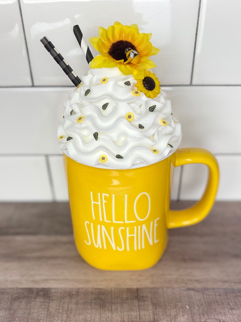 May include: Yellow ceramic mug with the text "Hello Sunshine" in white. The mug is topped with white whipped cream, yellow and black sunflower decorations, and black and white striped straws.