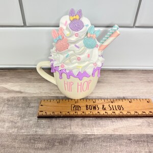 K&ouml;nnte beinhalten: Eine wei&szlig;e Keramiktasse mit lila Glasur und den Worten "Hip Hop" auf der Vorderseite. Die Tasse ist gef&uuml;llt mit wei&szlig;er Schlagsahne, pastellfarbenen Streuseln und drei Hasenfiguren mit Schleifen. Ein gestreifter Strohhalm ragt aus der Oberseite.