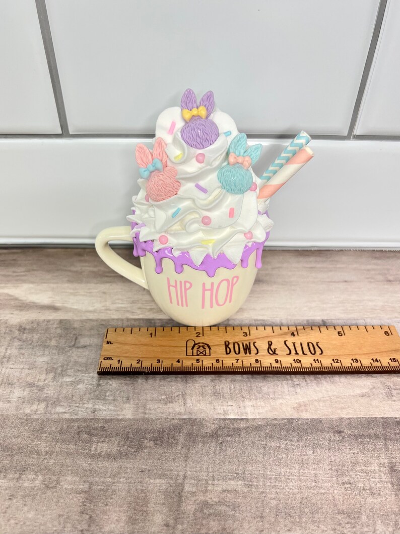 K&ouml;nnte beinhalten: Eine wei&szlig;e Keramiktasse mit lila Glasur und den Worten "Hip Hop" auf der Vorderseite. Die Tasse ist gef&uuml;llt mit wei&szlig;er Schlagsahne, pastellfarbenen Streuseln und drei Hasenfiguren mit Schleifen. Ein gestreifter Strohhalm ragt aus der Oberseite.