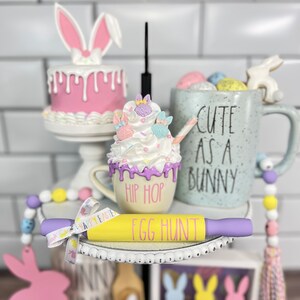 K&ouml;nnte beinhalten: Ein dreistufiges Tablett, das f&uuml;r Ostern mit einem rosa Hasenkuchen, einer Tasse mit Schlagsahne und Streuseln, einer Tasse mit der Aufschrift "Cute as a Bunny" und einem gelben Nudelholz mit der Aufschrift "Egg Hunt" dekoriert ist.