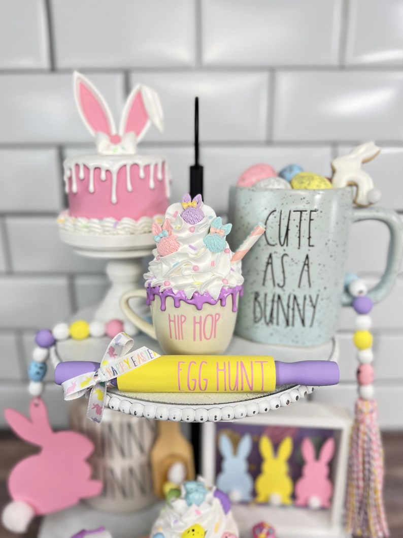 K&ouml;nnte beinhalten: Ein dreistufiges Tablett, das f&uuml;r Ostern mit einem rosa Hasenkuchen, einer Tasse mit Schlagsahne und Streuseln, einer Tasse mit der Aufschrift "Cute as a Bunny" und einem gelben Nudelholz mit der Aufschrift "Egg Hunt" dekoriert ist.