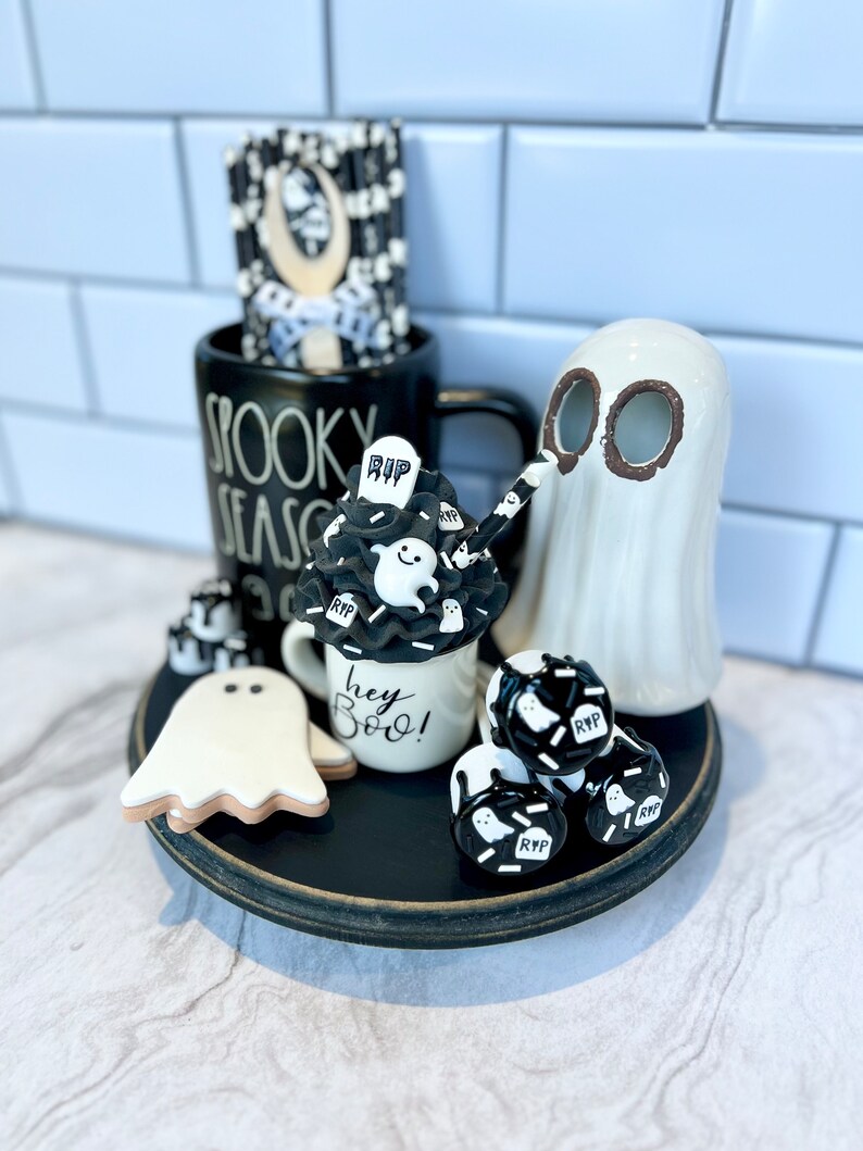 May include: A black and white Halloween themed tiered tray with a ghost, a mug that says "Spooky Season", a mug that says "Hey Boo!" and a variety of black and white ghost-shaped decorations.