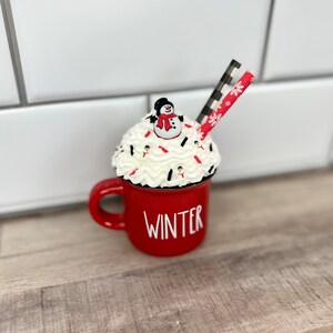Op de afbeelding: Rode keramische mok met witte letters die "WINTER" zeggen. De mok is gevuld met slagroom en rode en witte sprinkles. Een klein sneeuwpopfiguurtje zit bovenop de slagroom. Een rood-wit gestreepte rietje steekt uit de slagroom.