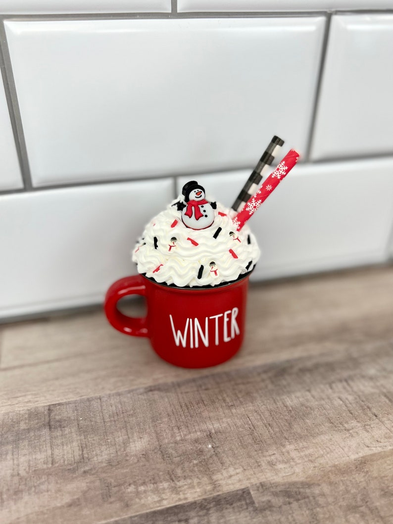 Op de afbeelding: Rode keramische mok met witte letters die "WINTER" zeggen. De mok is gevuld met slagroom en rode en witte sprinkles. Een klein sneeuwpopfiguurtje zit bovenop de slagroom. Een rood-wit gestreepte rietje steekt uit de slagroom.