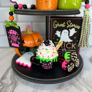 May include: A tiered tray decorated for Halloween with a black and white ghost, a pumpkin, a green mug with the text "Boo", a black mug with the text "I'm a haunt mess", and a sign that says "Ghost Stories Trick or Treat".