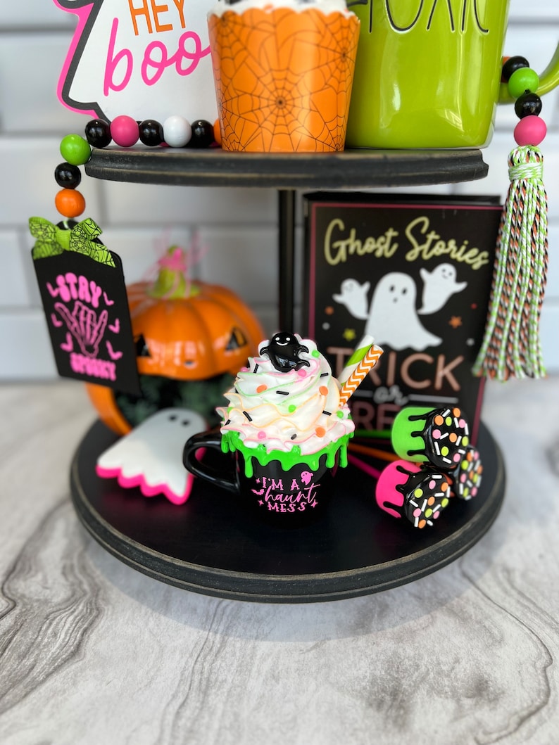 May include: A tiered tray decorated for Halloween with a black and white ghost, a pumpkin, a green mug with the text "Boo", a black mug with the text "I'm a haunt mess", and a sign that says "Ghost Stories Trick or Treat".