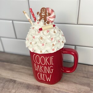 May include: Red ceramic mug with a white whipped cream topping, a gingerbread man, a candy cane, and a rolling pin. The mug says "Cookie Baking Crew".