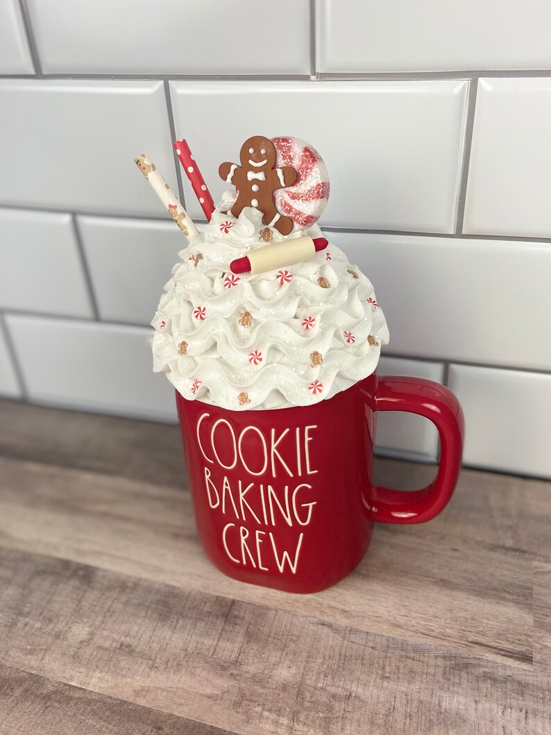 May include: Red ceramic mug with a white whipped cream topping, a gingerbread man, a candy cane, and a rolling pin. The mug says "Cookie Baking Crew".