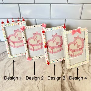 May include: Four white picture frames with a frosting-like design and colorful sprinkles. Each frame has a pink heart with the text "Bows & Silos" inside. The frames are labeled "Design 1", "Design 2", "Design 3", and "Design 4".