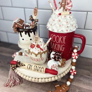 May include: A tiered tray decorated for the holidays with a red mug that says "Cookie Baking Crew", a gingerbread mug with whipped cream and chocolate drizzle, a white mug with a gingerbread house and chocolate drizzle, a rolling pin that says "Oh Snap!" and a gingerbread man ornament.