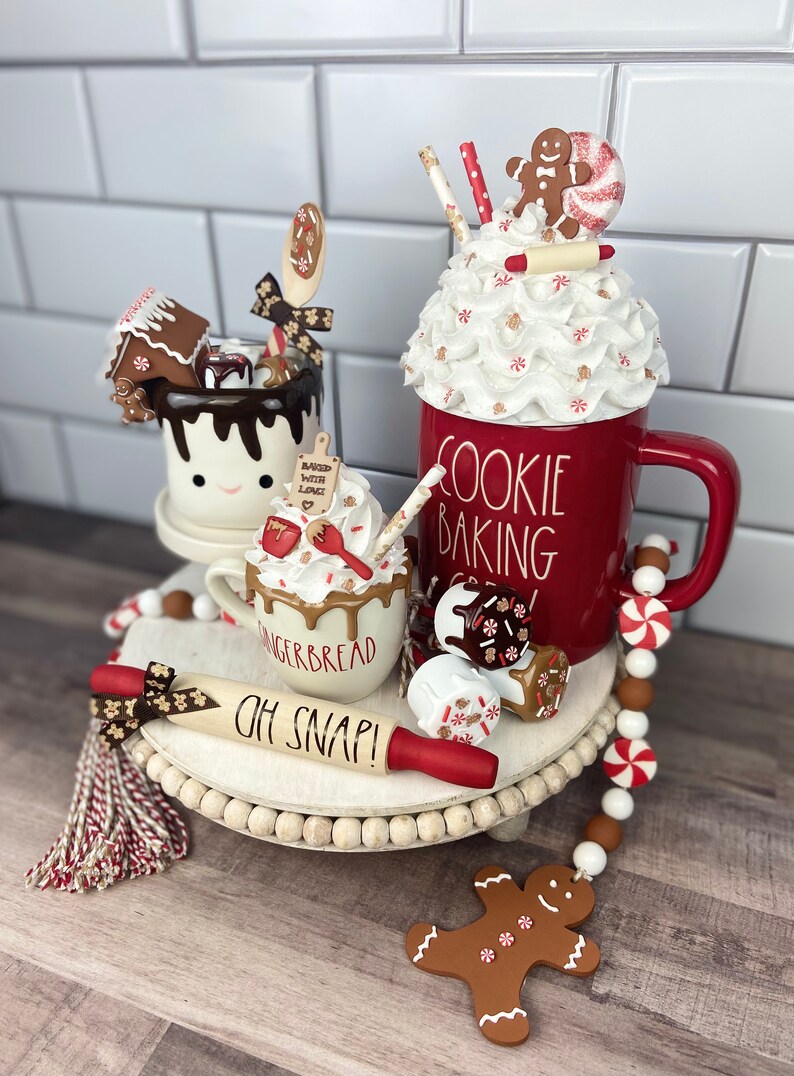 May include: A tiered tray decorated for the holidays with a red mug that says "Cookie Baking Crew", a gingerbread mug with whipped cream and chocolate drizzle, a white mug with a gingerbread house and chocolate drizzle, a rolling pin that says "Oh Snap!" and a gingerbread man ornament.