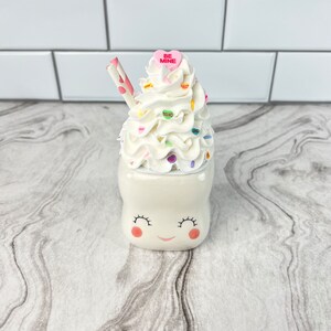 May include: White ceramic mug with a smiling face and pink cheeks. The mug is topped with whipped cream, sprinkles, and a pink candy heart that says "Be Mine". A striped straw sticks out of the whipped cream.