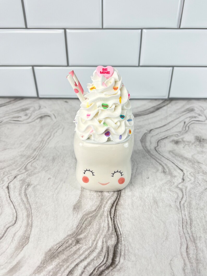 May include: White ceramic mug with a smiling face and pink cheeks. The mug is topped with whipped cream, sprinkles, and a pink candy heart that says "Be Mine". A striped straw sticks out of the whipped cream.