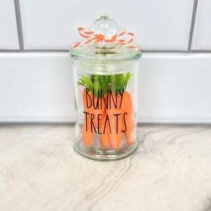 May include: A clear glass jar with a lid tied with a peach and white ribbon. The jar contains carrot-shaped decorations and the text "BUNNY TREATS" is printed on the front.