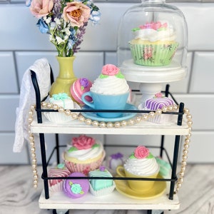 May include: A white two-tiered rolling cart with black metal accents, decorated with pastel-colored cupcakes, macarons, and other pastries. A bouquet of pink and blue flowers sits in a yellow vase on the top shelf.