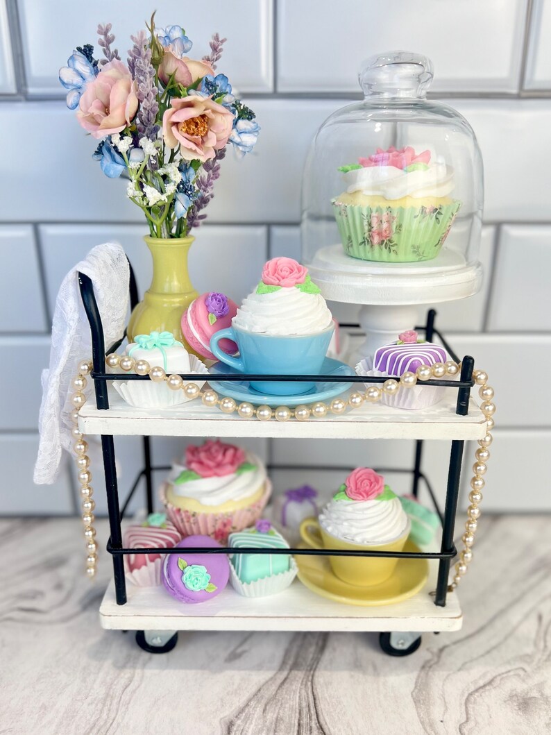 May include: A white two-tiered rolling cart with black metal accents, decorated with pastel-colored cupcakes, macarons, and other pastries. A bouquet of pink and blue flowers sits in a yellow vase on the top shelf.