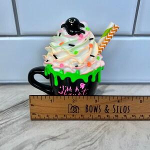 May include: A black ceramic mug with green and white frosting, topped with a black ghost and sprinkles. The mug reads "I'm a Ghoul".
