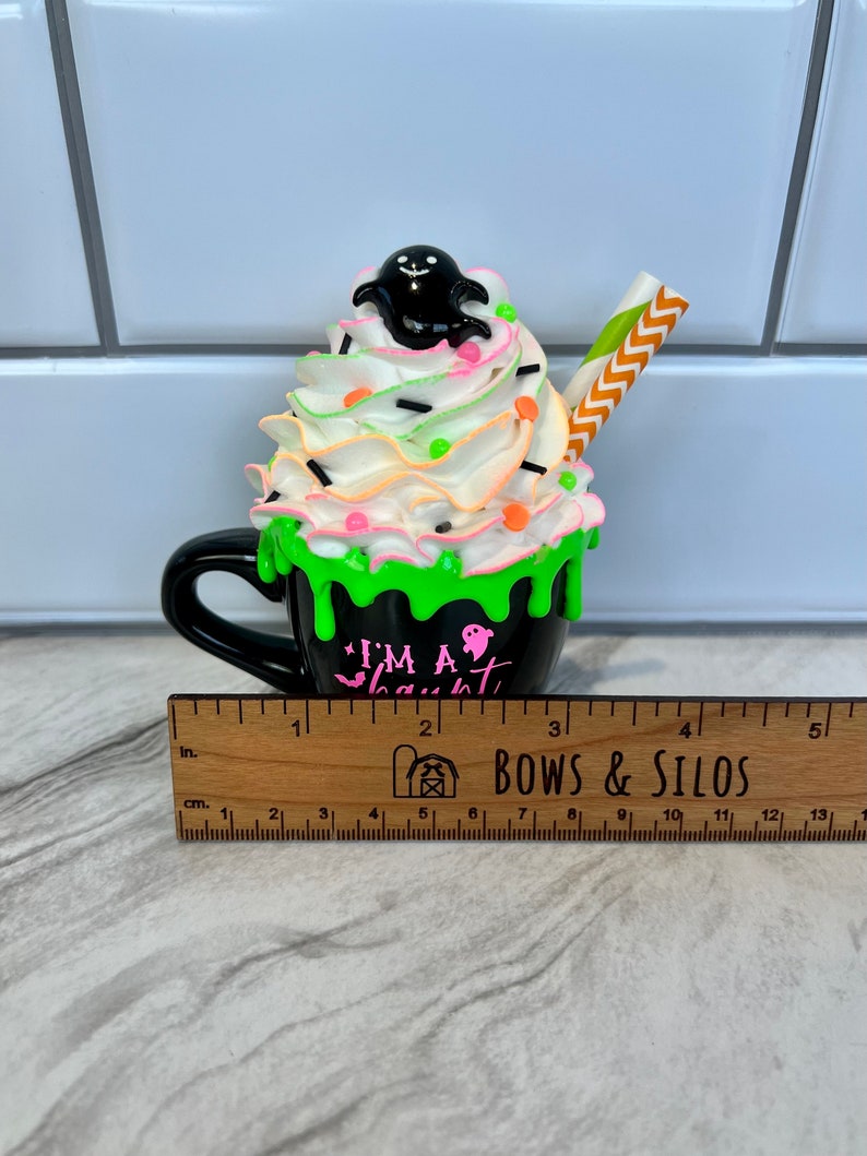 May include: A black ceramic mug with green and white frosting, topped with a black ghost and sprinkles. The mug reads "I'm a Ghoul".