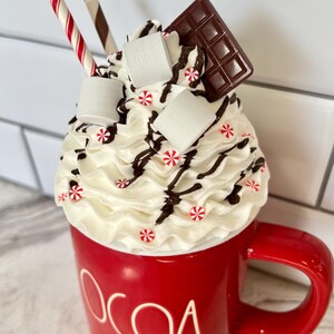 May include: A red mug with the word "COCOA" on the side, filled with whipped cream, chocolate shavings, marshmallows, and candy canes.