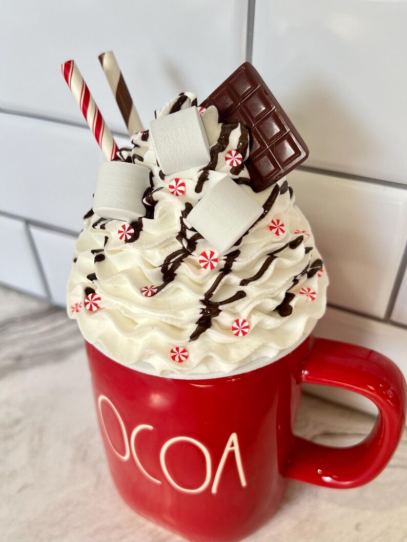 May include: A red mug with the word "COCOA" on the side, filled with whipped cream, chocolate shavings, marshmallows, and candy canes.