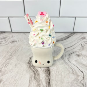 May include: A white ceramic mug with a smiling face, topped with whipped cream and colorful sprinkles. The mug has a pink heart candy with the words "BE MINE" on it.