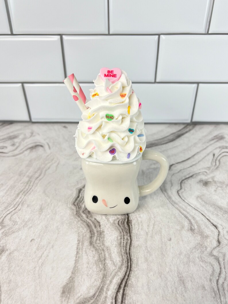 May include: A white ceramic mug with a smiling face, topped with whipped cream and colorful sprinkles. The mug has a pink heart candy with the words "BE MINE" on it.