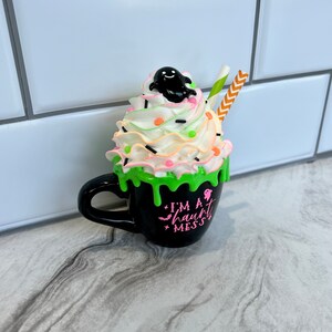 May include: A black coffee mug with a green slime drip and a white whipped cream topping. The mug has a ghost on top of the whipped cream and says "I'm a haunt mess".