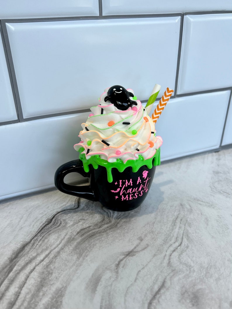 May include: A black coffee mug with a green slime drip and a white whipped cream topping. The mug has a ghost on top of the whipped cream and says "I'm a haunt mess".