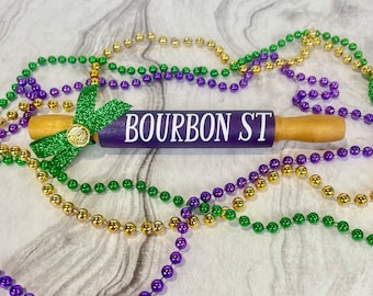 Puede incluir: Rodillo de amasar morado con el texto "BOURBON ST" en letras blancas. El rodillo está decorado con un lazo de purpurina verde y rodeado de cuentas moradas, verdes y doradas.