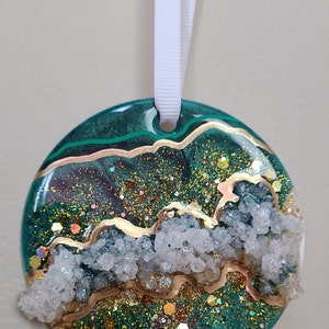 May include: A round green and gold glitter ornament with white crystal accents. The ornament has a white ribbon attached for hanging.