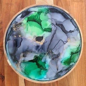 May include: A round wooden tray with a green, gray, and gold abstract design. The design is created with resin and alcohol inks, creating a marbled effect.