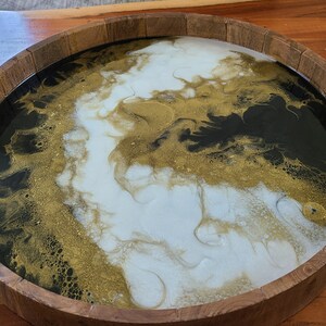May include: A round wooden tray with a black, white and gold epoxy resin design. The resin has a marbled effect with swirls of black, white and gold.
