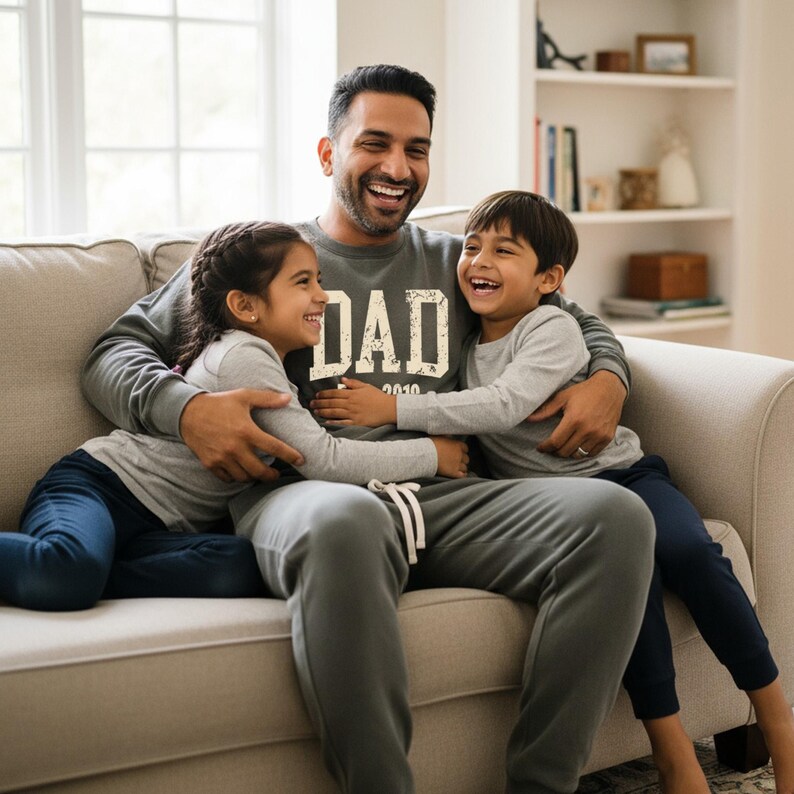 May include: A man wearing a gray sweatshirt with the word "DAD" printed on it, hugs two children on a beige couch. The children and the man are smiling. The man is wearing gray sweatpants. The children are wearing gray long-sleeve shirts and blue pants.