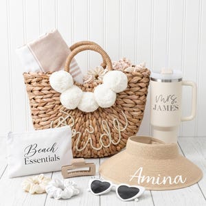 May include: A woven straw tote bag with white pom-poms and the word Bride, a beige tumbler with the text Mrs. James, a white pouch labeled Beach Essentials, a beige sun visor with the name Amina, heart-shaped sunglasses, and two hair scrunchies.