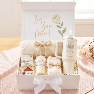 May include: A white gift box filled with a cream-colored blanket, a tumbler with "Best Mom Ever" text, a chocolate bar, a candle, and a washcloth. The box lid has "Love You Mom!" text and a floral design. A white bow adorns the box.