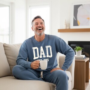 May include: A blue sweatshirt and matching sweatpants set with the word "DAD" in white block letters and "EST. 1985" underneath. The person is holding a white mug and sitting on a beige couch.