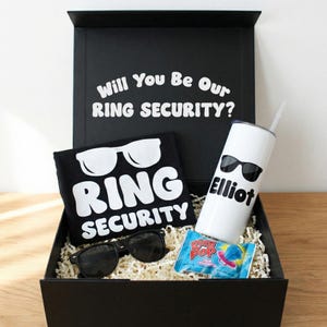 May include: A black gift box with the text "Will You Be Our RING SECURITY?" contains a black t-shirt, sunglasses, a white tumbler, and a blue raspberry Ring Pop. The t-shirt and tumbler have sunglasses and the words "RING SECURITY" and "Elliot" printed on them.
