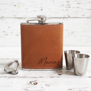 K&ouml;nnte beinhalten: Eine braune Lederflasche mit dem Namen "Marcus" auf der Vorderseite eingraviert. Zwei kleine Schnapsgl&auml;ser aus Edelstahl und ein silberner Trichter befinden sich neben der Flasche. Die Flasche hat einen silbernen Deckel.