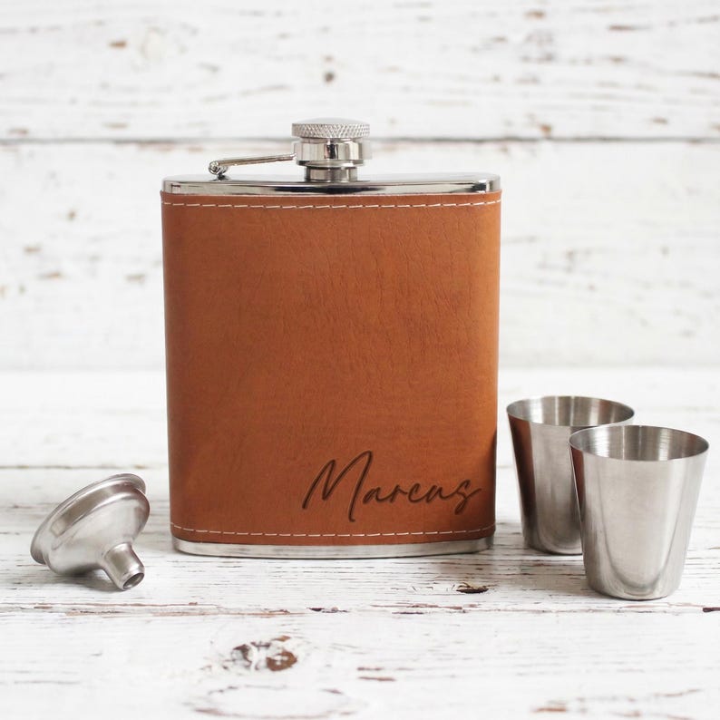 K&ouml;nnte beinhalten: Eine braune Lederflasche mit dem Namen "Marcus" auf der Vorderseite eingraviert. Zwei kleine Schnapsgl&auml;ser aus Edelstahl und ein silberner Trichter befinden sich neben der Flasche. Die Flasche hat einen silbernen Deckel.
