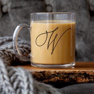 May include: A clear glass mug with a black cursive "JW" monogram on the side, filled with a light brown beverage. The mug is sitting on a wooden surface.