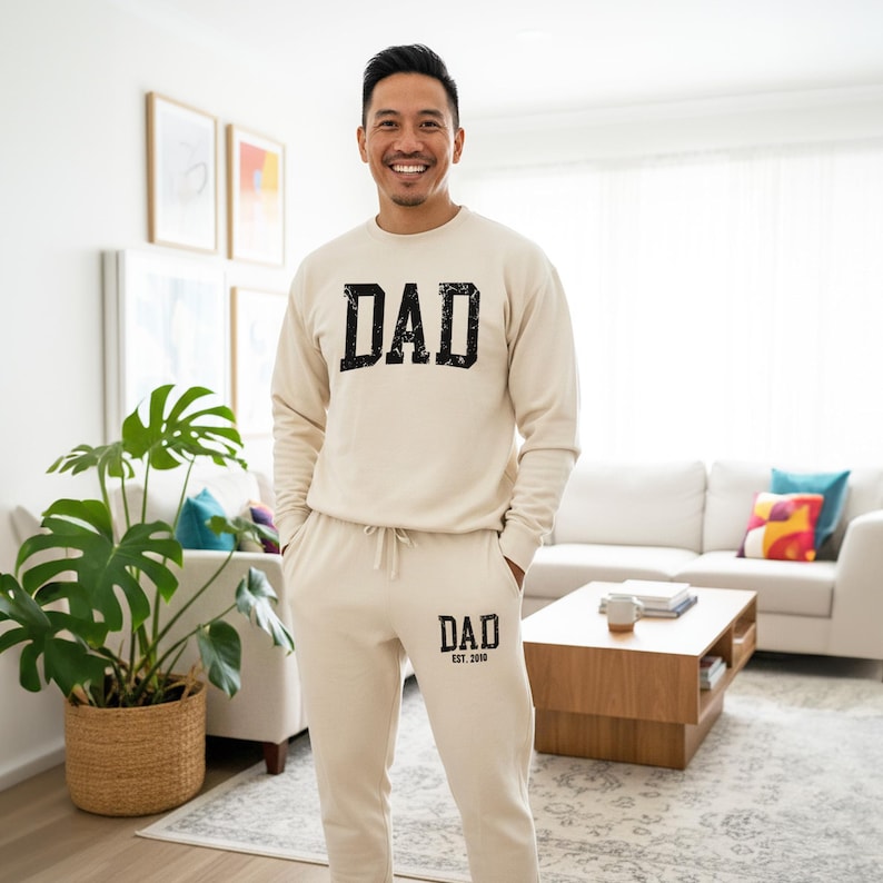 May include: A beige sweatshirt and matching sweatpants set. The word "DAD" is printed in a distressed black font on the sweatshirt and sweatpants. The sweatpants also have "EST. 2018" printed below the word "DAD".
