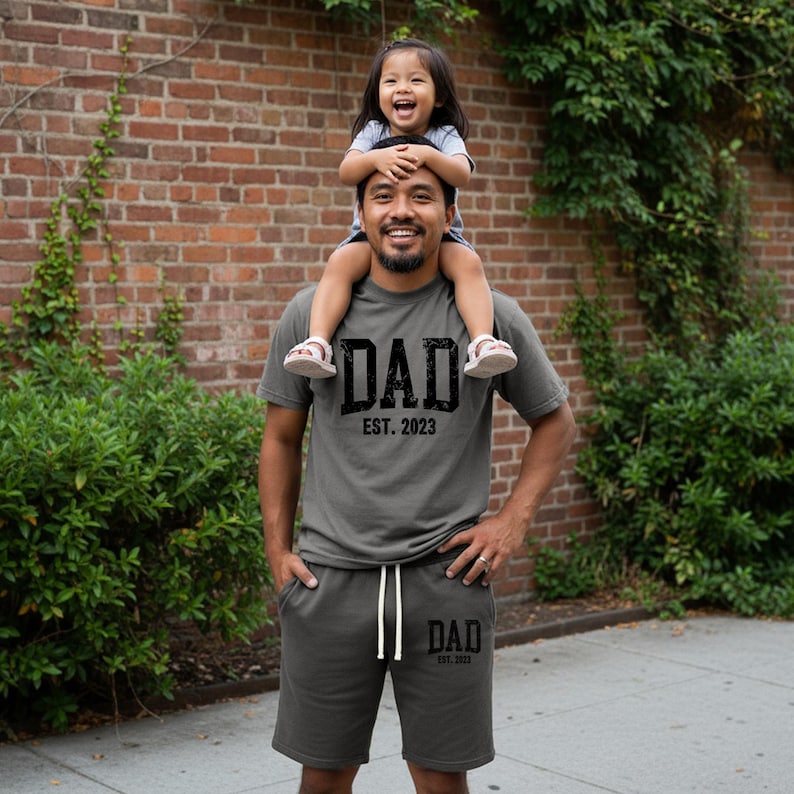 May include: A matching gray t-shirt and shorts set with the word "DAD EST. 2023" printed in black. A person is wearing the set, with a child sitting on their shoulders. The background is a brick wall and green foliage.