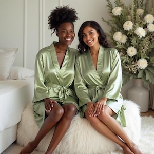 May include: Two people wearing matching sage green satin robes with a tie belt. They are sitting on a fluffy white rug and smiling at the camera.