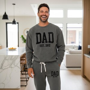 May include: A gray sweatshirt and matching sweatpants set with the word "DAD" and "EST. 2012" printed in black. The sweatpants have a white drawstring. The person in the image is smiling.
