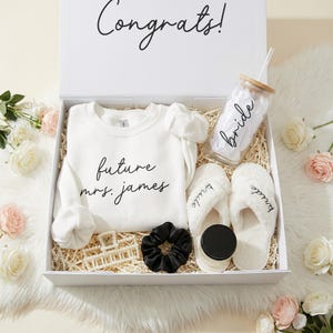 Puede incluir: Una caja de regalo blanca con la palabra "Congrats!" en la tapa, que contiene una sudadera blanca con "future mrs. james" impresa, un vaso de vidrio con "bride" impreso, zapatillas blancas con "bride" bordado y otros accesorios.