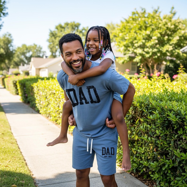 May include: A man and a child are outside on a sunny day. The man is wearing a blue t-shirt and shorts set with the word "DAD" printed on the front. The child is riding on the man's back and smiling.
