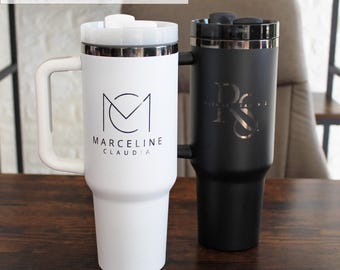 May include: Two stainless steel travel mugs with lids. The mug on the left is white with a black logo that says "Marceline Claudia". The mug on the right is black with a silver logo that says "R.S.". Both mugs have a handle.
