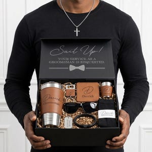 May include: A black gift box containing a tumbler, wallet, shot glass, sunglasses, bracelet, keychain, and socks. The box lid has the text "Suit Up! Your service as a groomsman is requested."