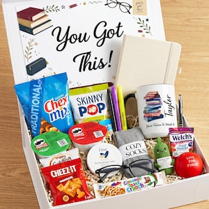 May include: A white gift box filled with snacks, a notebook, a mug, pens, glasses, and a stress ball. The box lid has the text "You Got This!" and a stack of books. The mug has a stack of books and the name "Taylor".